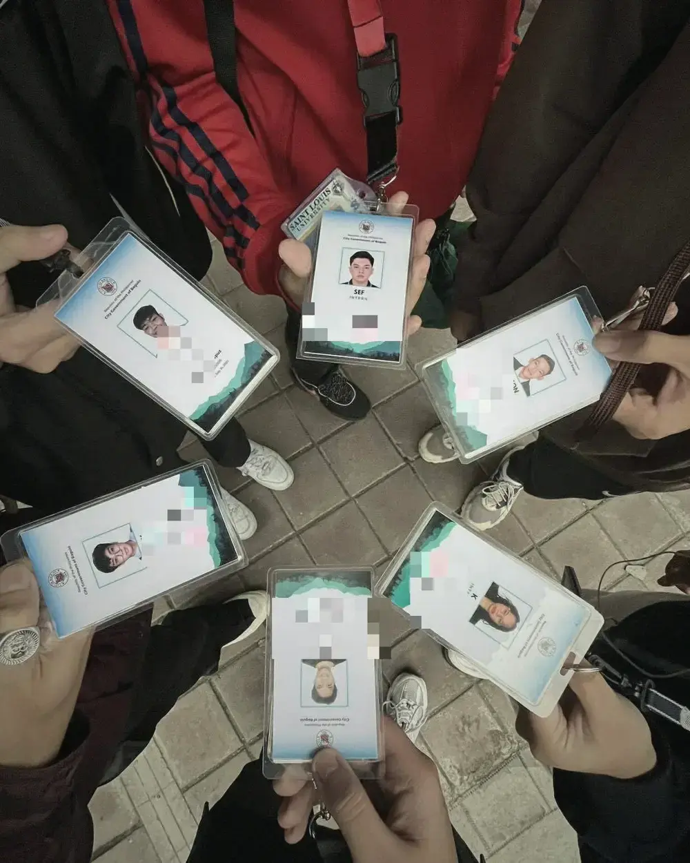 Hands holding City Government of Baguio intern ID cards out in a ring over a paved floor